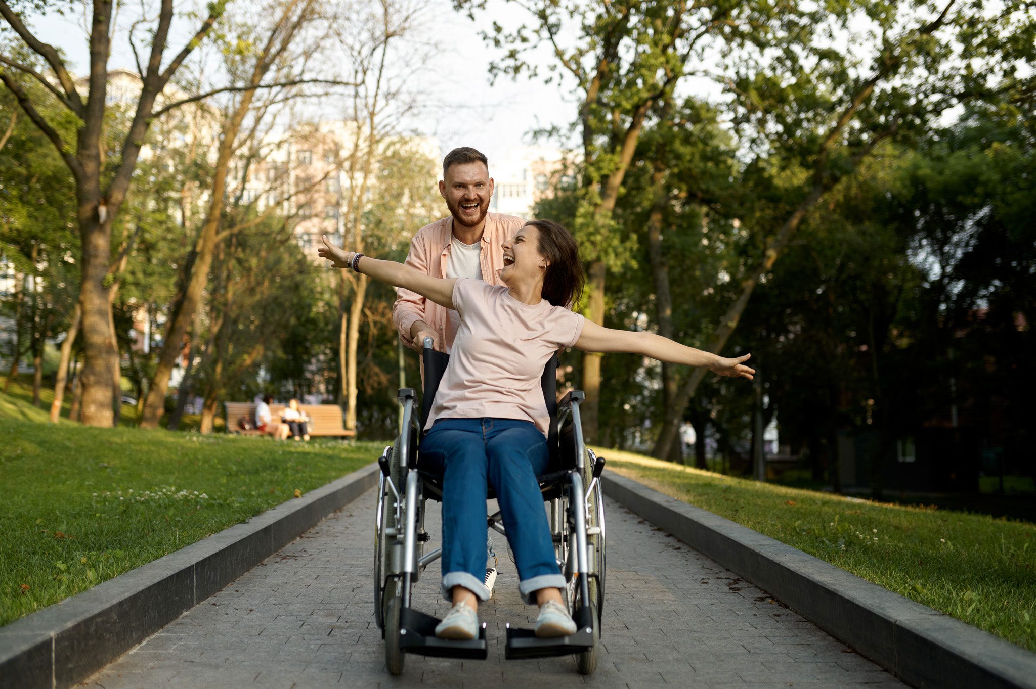 A man pushes a woman in a wheelchair along a park path; she smiles with arms outstretched while he walks behind her, also smiling. Trees and sunlight fill the background.
