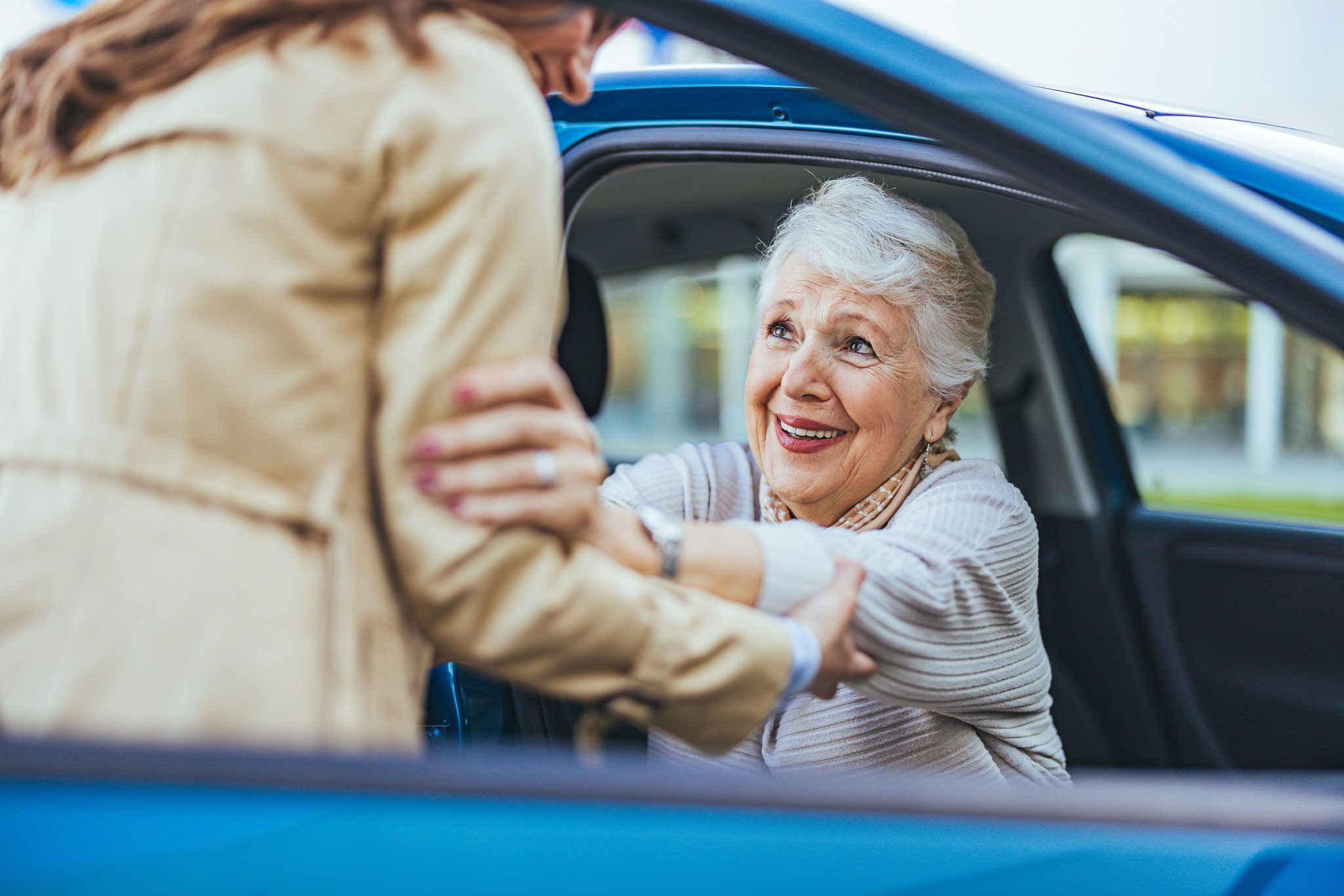 An elderly woman sitting in a car is being helped out by a younger person who is holding her arms and assisting her to stand.